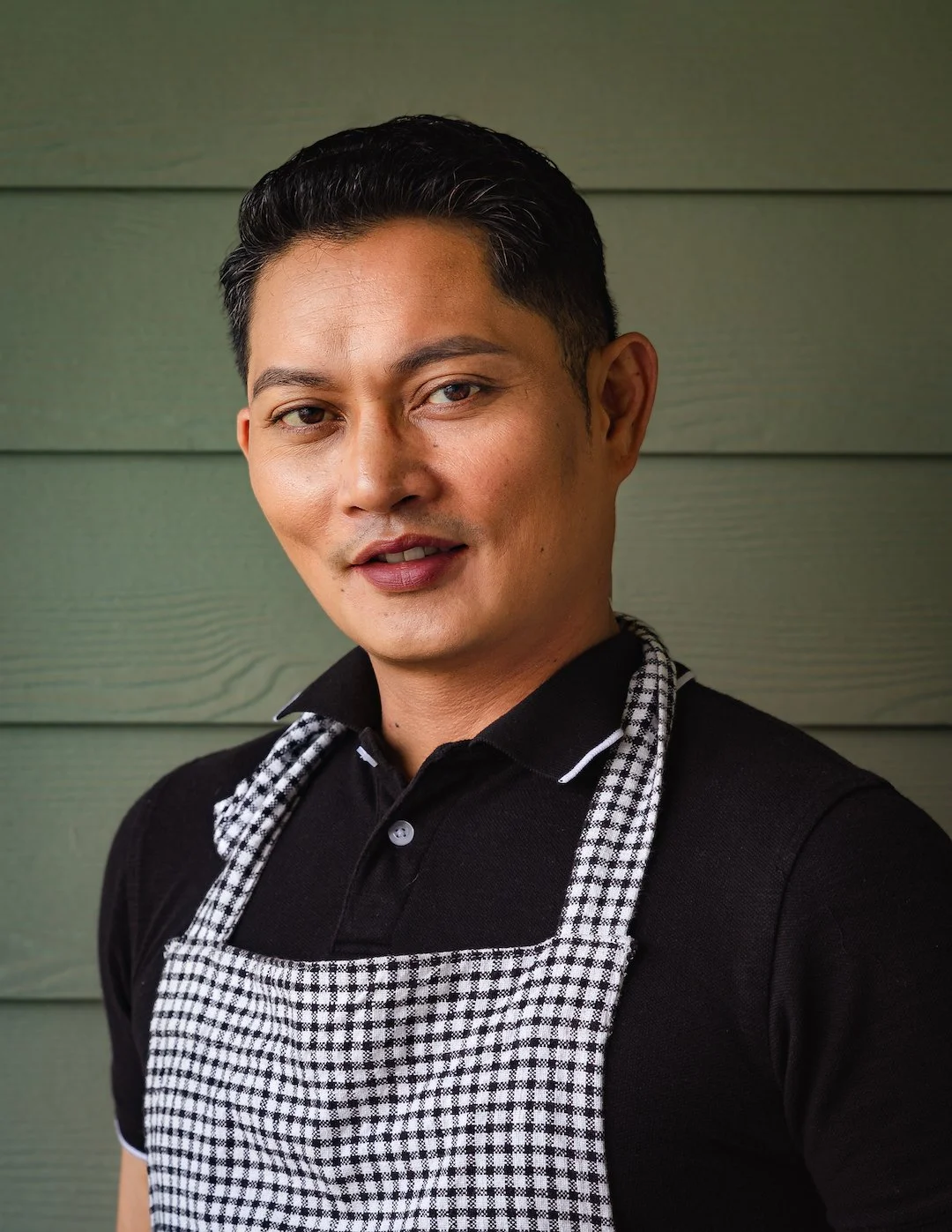 A man with dark hair and wearing a black shirt and checkered apron, standing against a green wooden wall.
