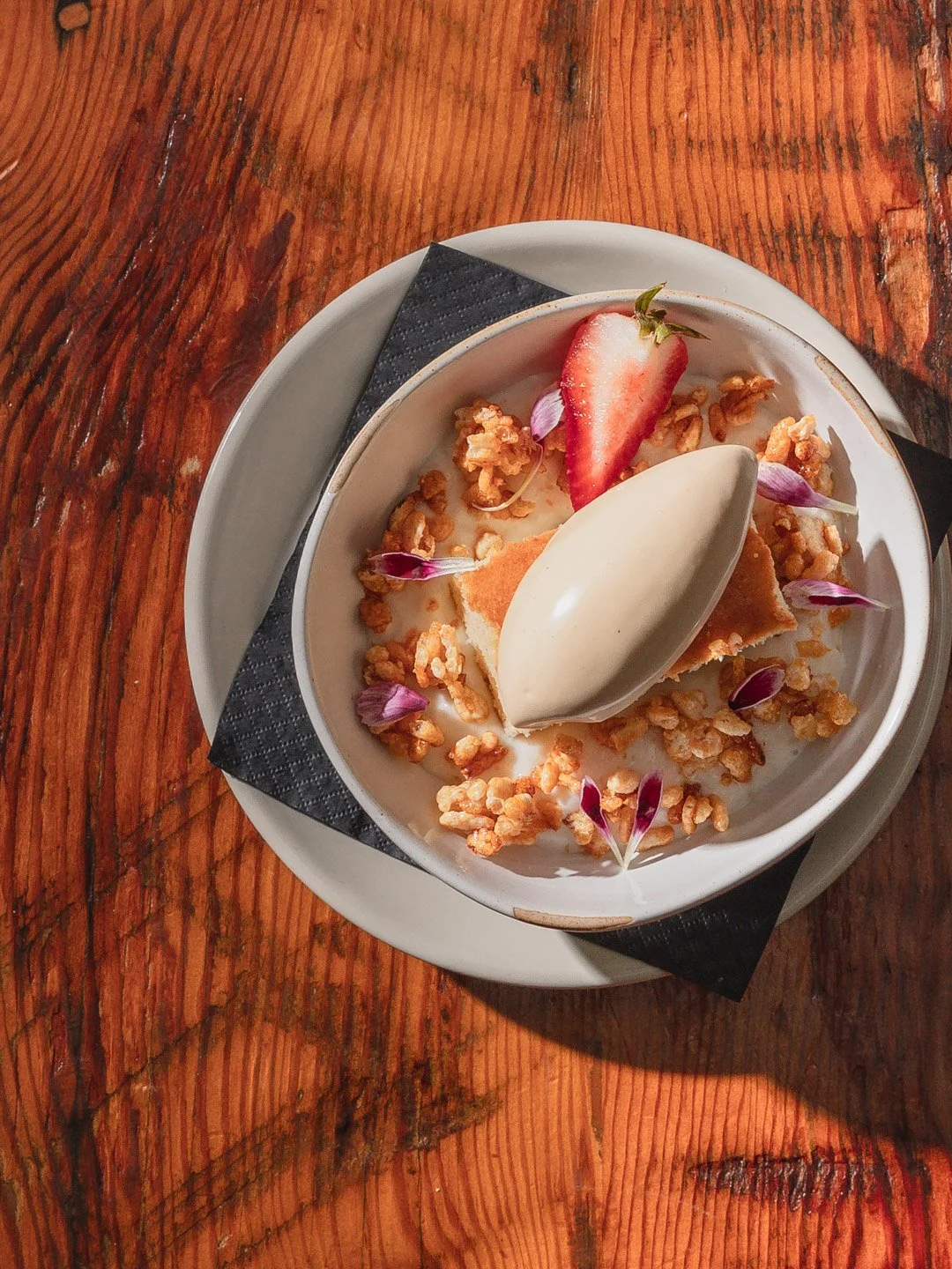 Dessert with a scoop of vanilla ice cream, fresh strawberry, edible flowers, and granola in a white bowl on a wooden table.