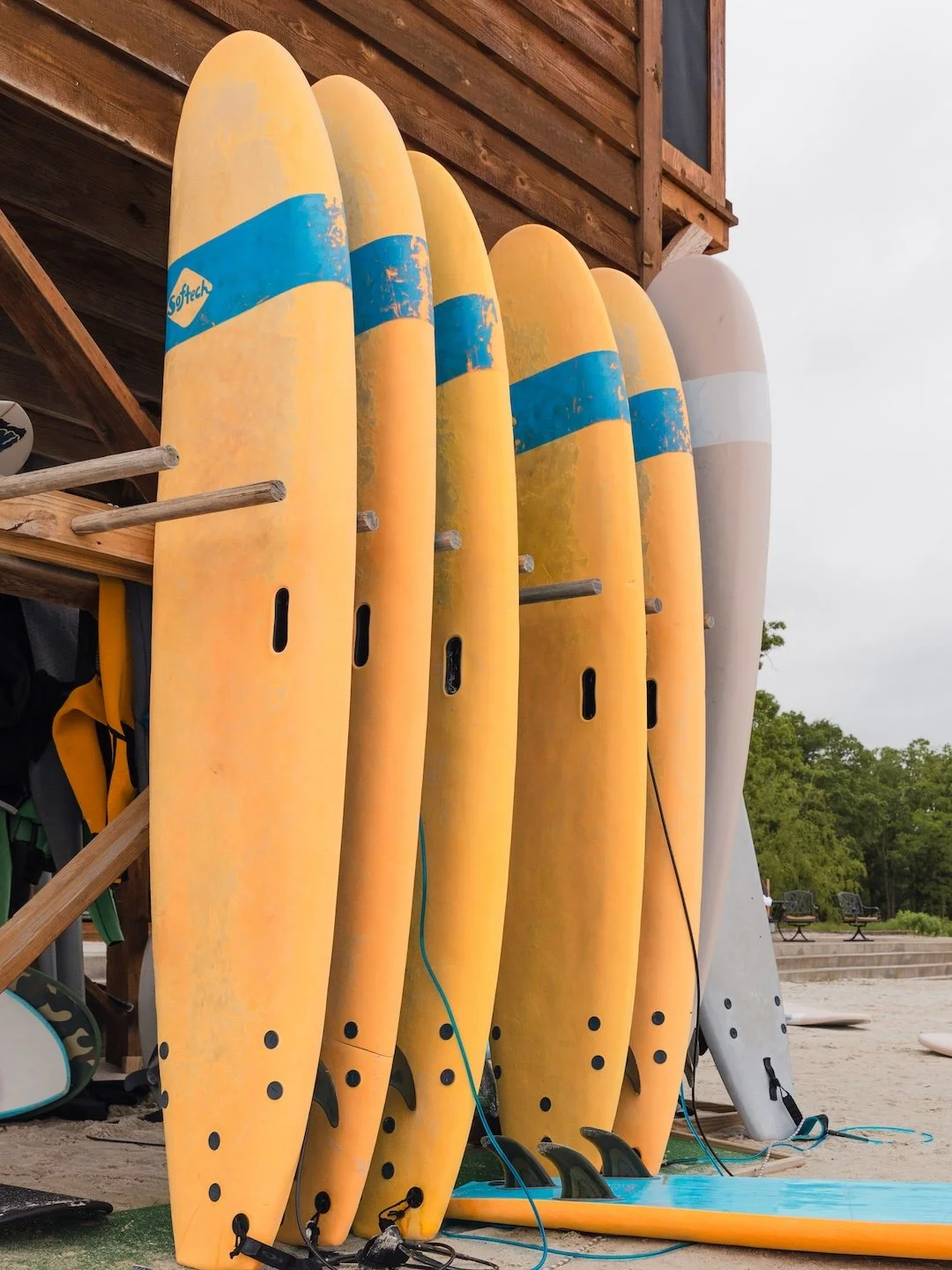 A group of surfboards lined up vertically against a wooden structure, with some surfboards showing signs of wear and bright colors, and the background includes a cloudy sky and trees.