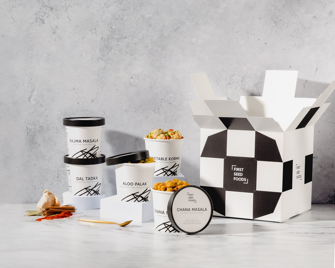 Collection of Indian food containers with spices, vegetables, and snacks on a white surface beside a black-and-white checkered food box.