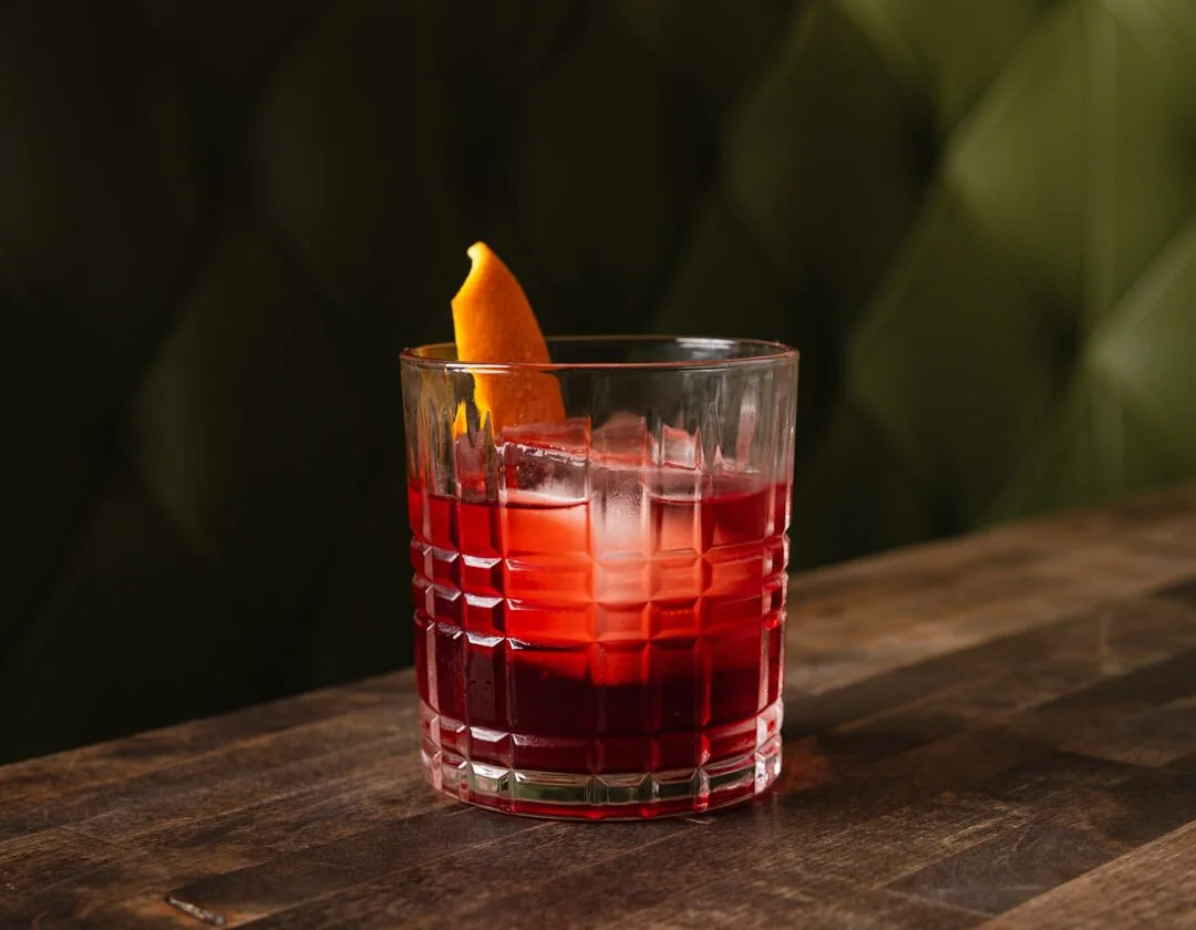 Red cocktail with ice cubes and an orange peel garnish in a cut-glass glass on a wooden surface.