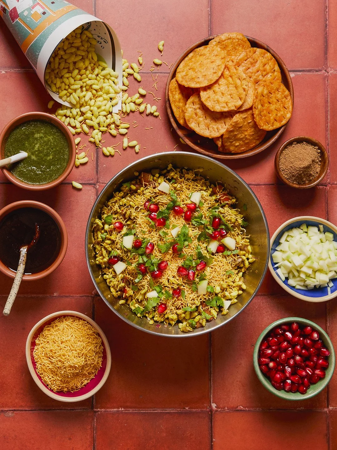 Spread of Indian snack foods and ingredients on a tiled surface, including a large bowl of chaat with pomegranate seeds, chopped onions, and cilantro, round potato chips, small bowls of syrup, chopped onions, ground spices, sev, and pomegranate seeds