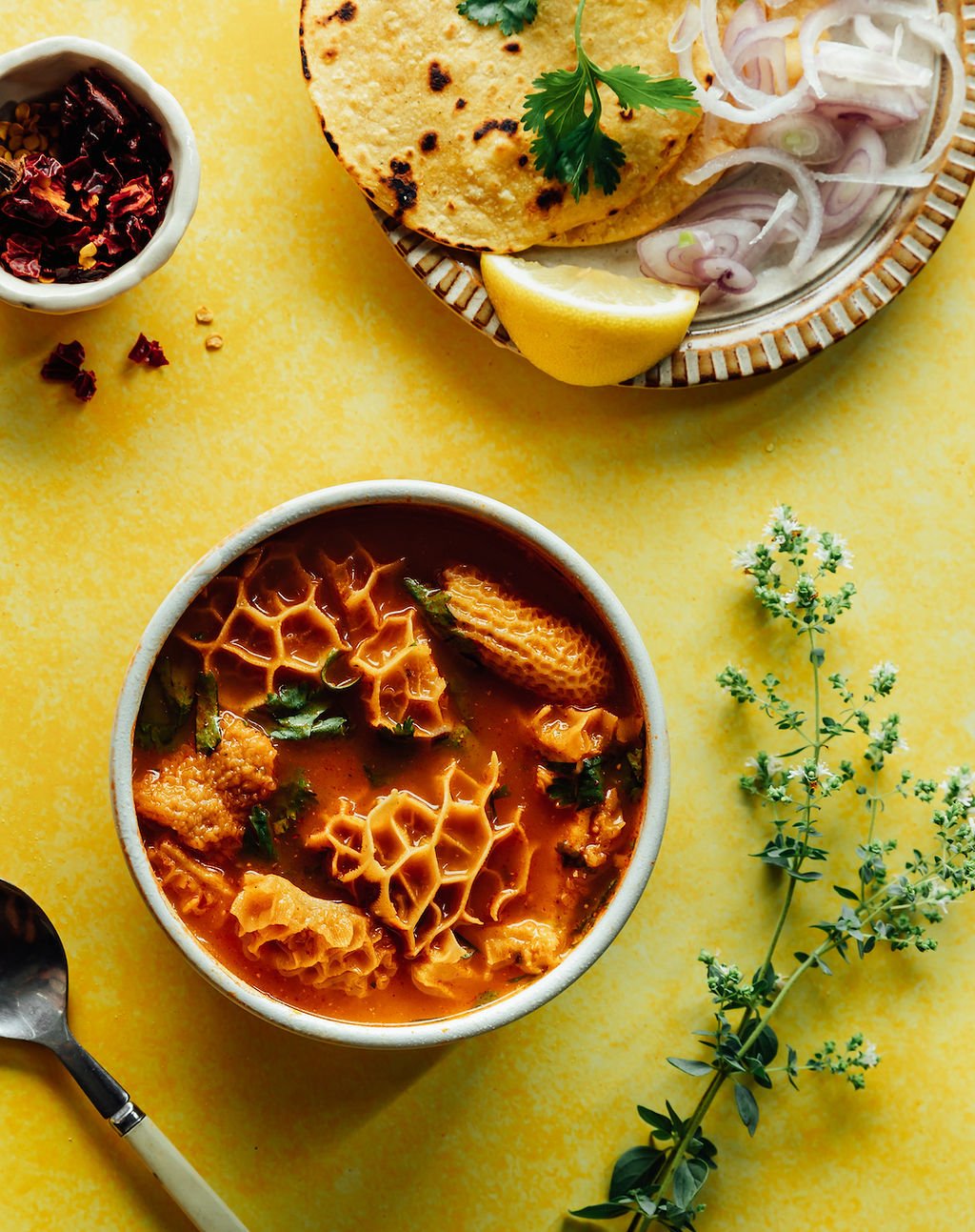 A bowl of Indian curry with cauliflower, herbs, and spices on a yellow surface, accompanied by a plate of naan bread, sliced onions, lemon wedge, and a small bowl of dried chili flakes.