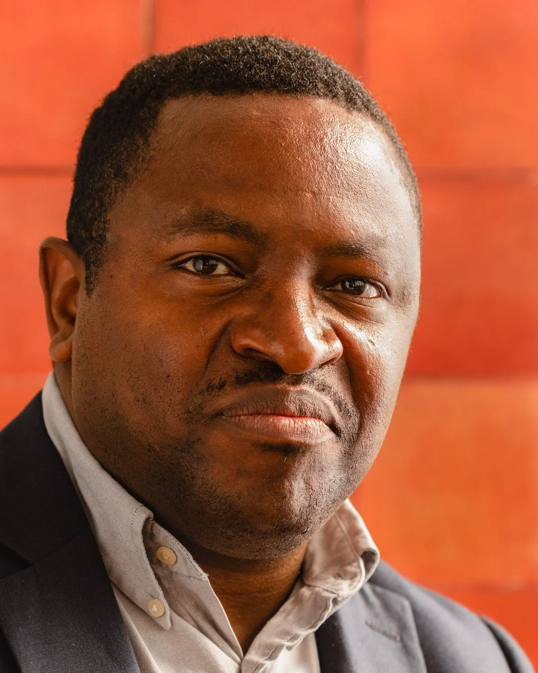A close-up portrait of a man with dark skin, short curly hair, wearing a light-colored button-up shirt and a dark blazer, against a soft orange background.