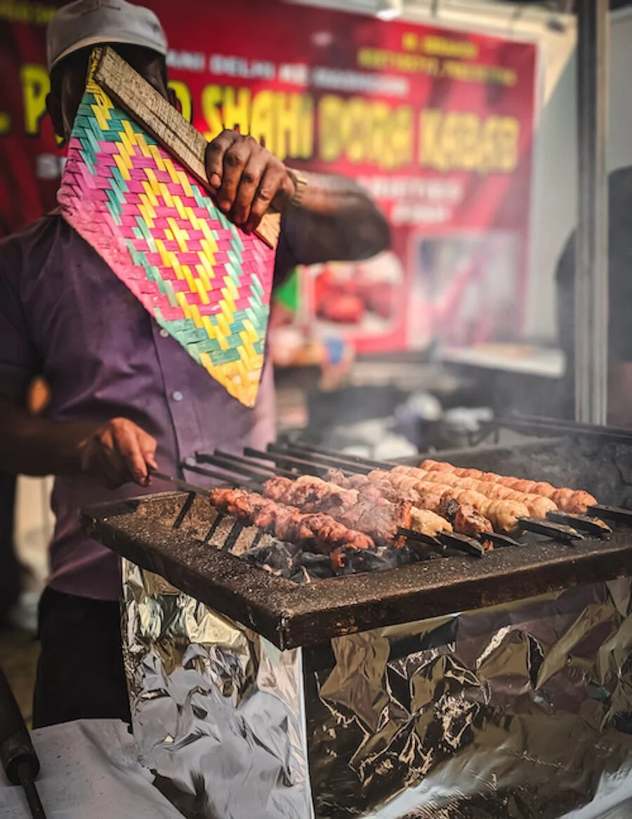 A street food vendor grilling skewers of meat over an open flame, holding a colorful woven mat in front of his face, with a red and yellow sign in the background.