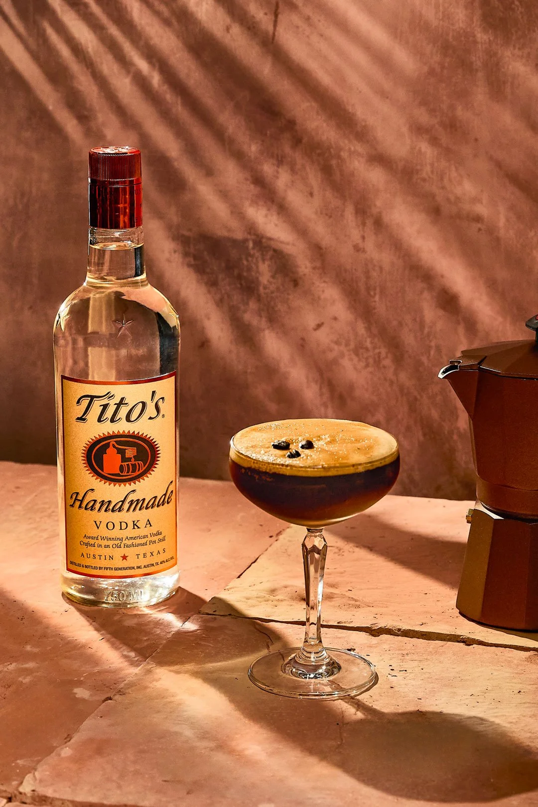 A bottle of Tito's Handmade Vodka, a cocktail glass filled with a dark drink garnished with coffee beans, and a small coffee maker on a pink marble surface with a pinkish textured background.