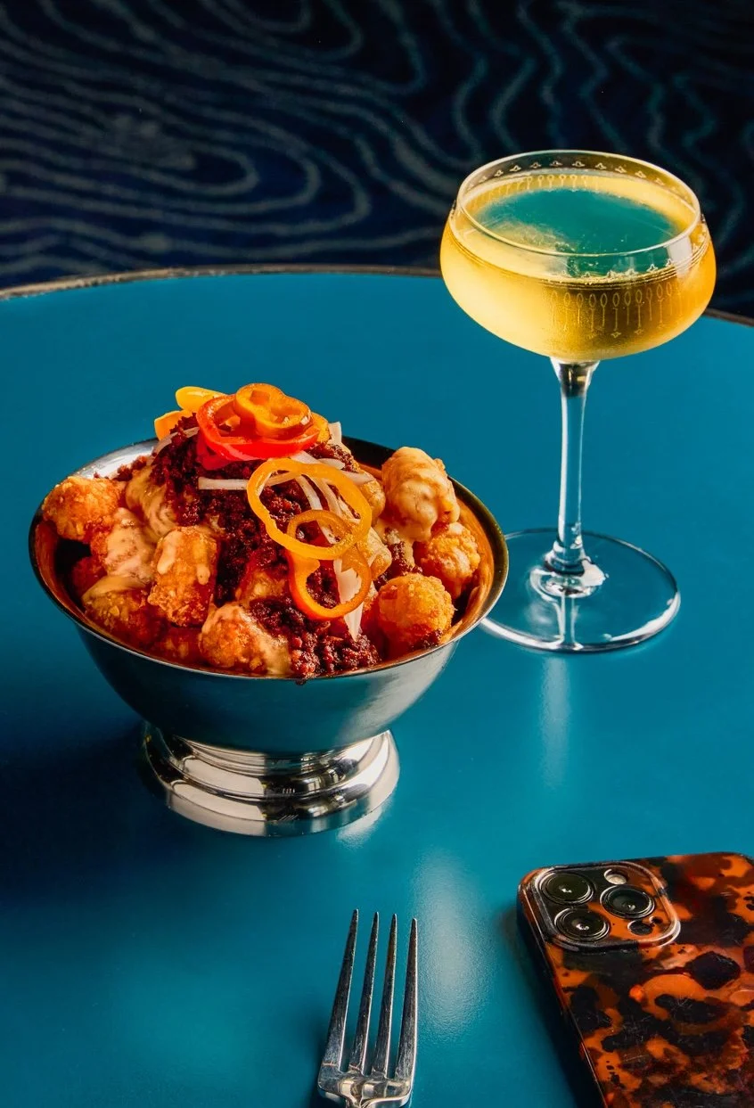 A bowl of loaded Asian-inspired chicken and vegetable dish topped with sliced peppers and fried wontons, alongside a glass of yellow cocktail on a blue table, with a fork and a smartphone with a leopard print case nearby.
