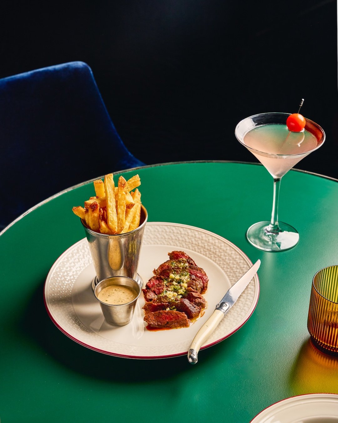 A plate of food with French fries, a small container of dipping sauce, and a sliced steak topped with herbs on a green table. To the right, there is a martini glass with a cocktail garnished with a cherry on a cocktail stick. An amber-colored glass i