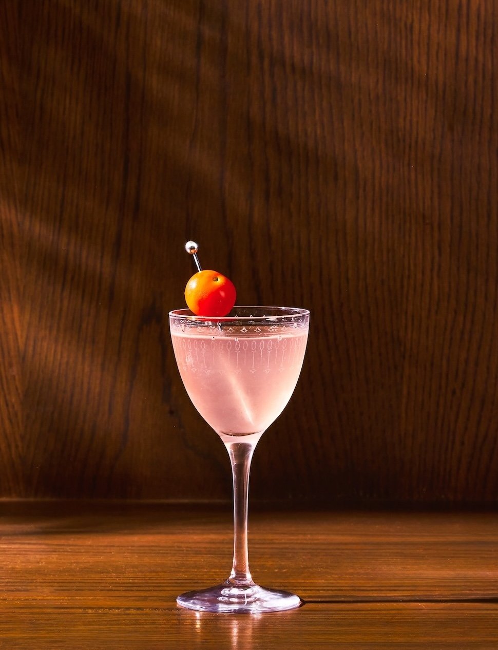 A pink cocktail in a tall, elegant glass garnished with a cherry and a decorative pick, set against a dark wooden background.