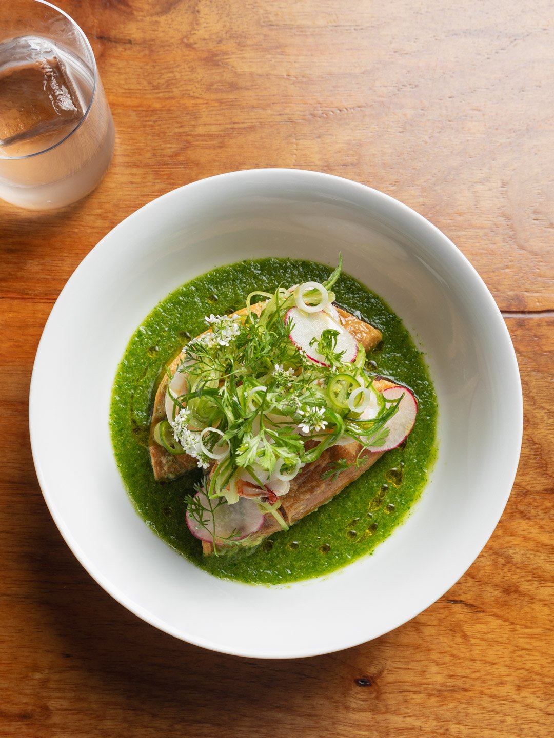 A white bowl with a green sauce, topped with sliced radishes, green onions, and herbs, with a piece of fish underneath. A glass of iced beverage is on a wooden table.