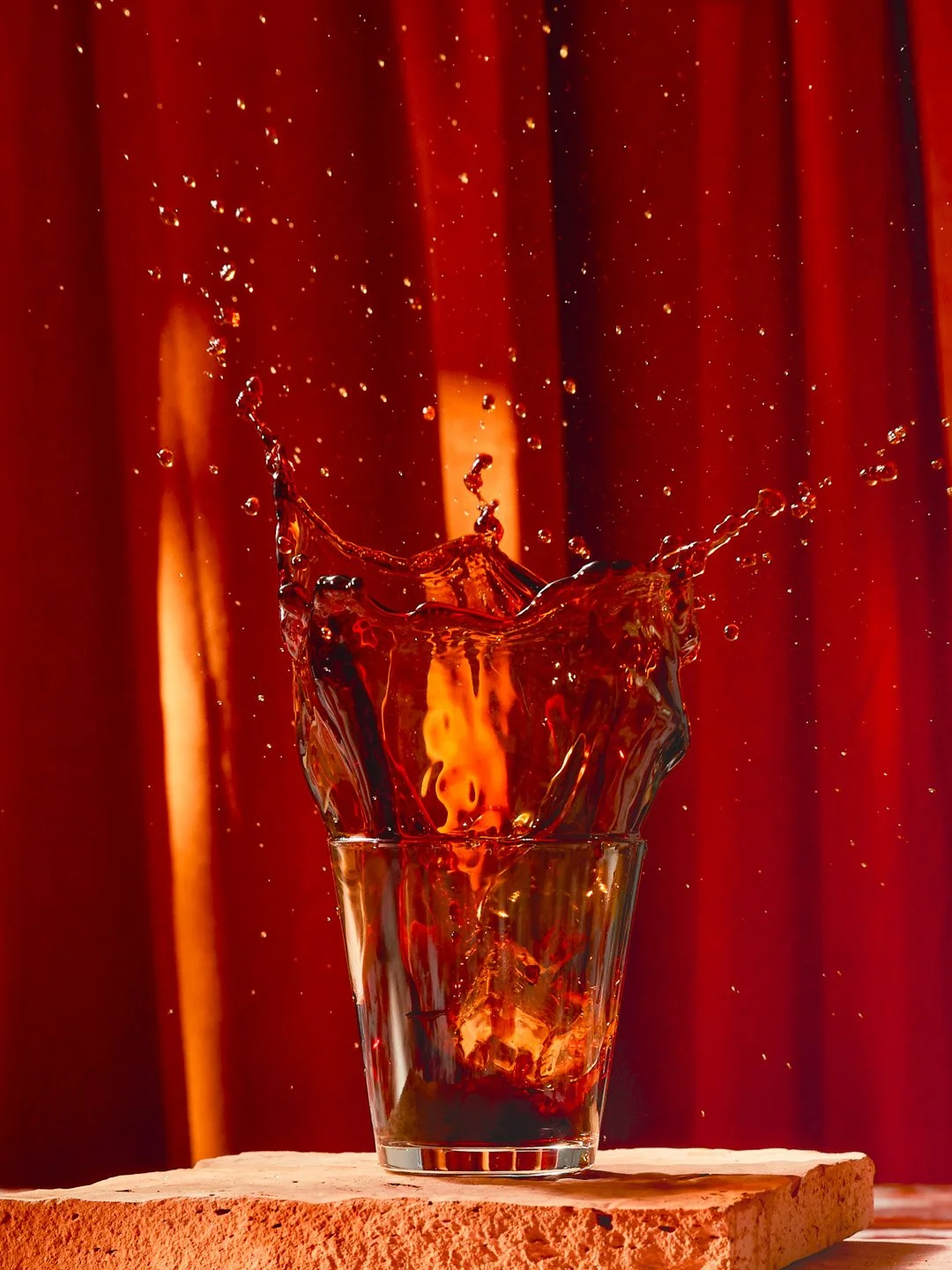 A splash of dark-colored liquid in a glass, captured mid-motion with droplets flying outward against a red curtain background.
