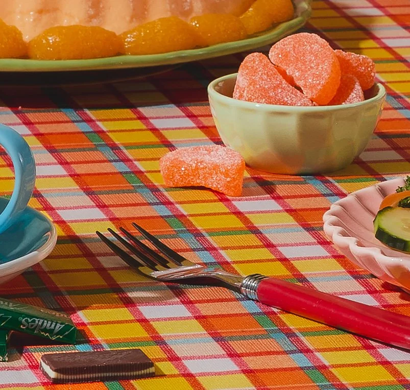 Colorful table setting with a checkered tablecloth, a fork with a red handle, a bowl of pink sugar-coated gumdrops, a plate with cucumber slices, and a larger plate with yellow and orange jelly slices.