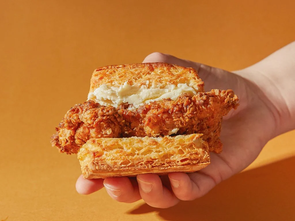 Hand holding a fried chicken sandwich with mashed potatoes and gravy inside, against an orange background.