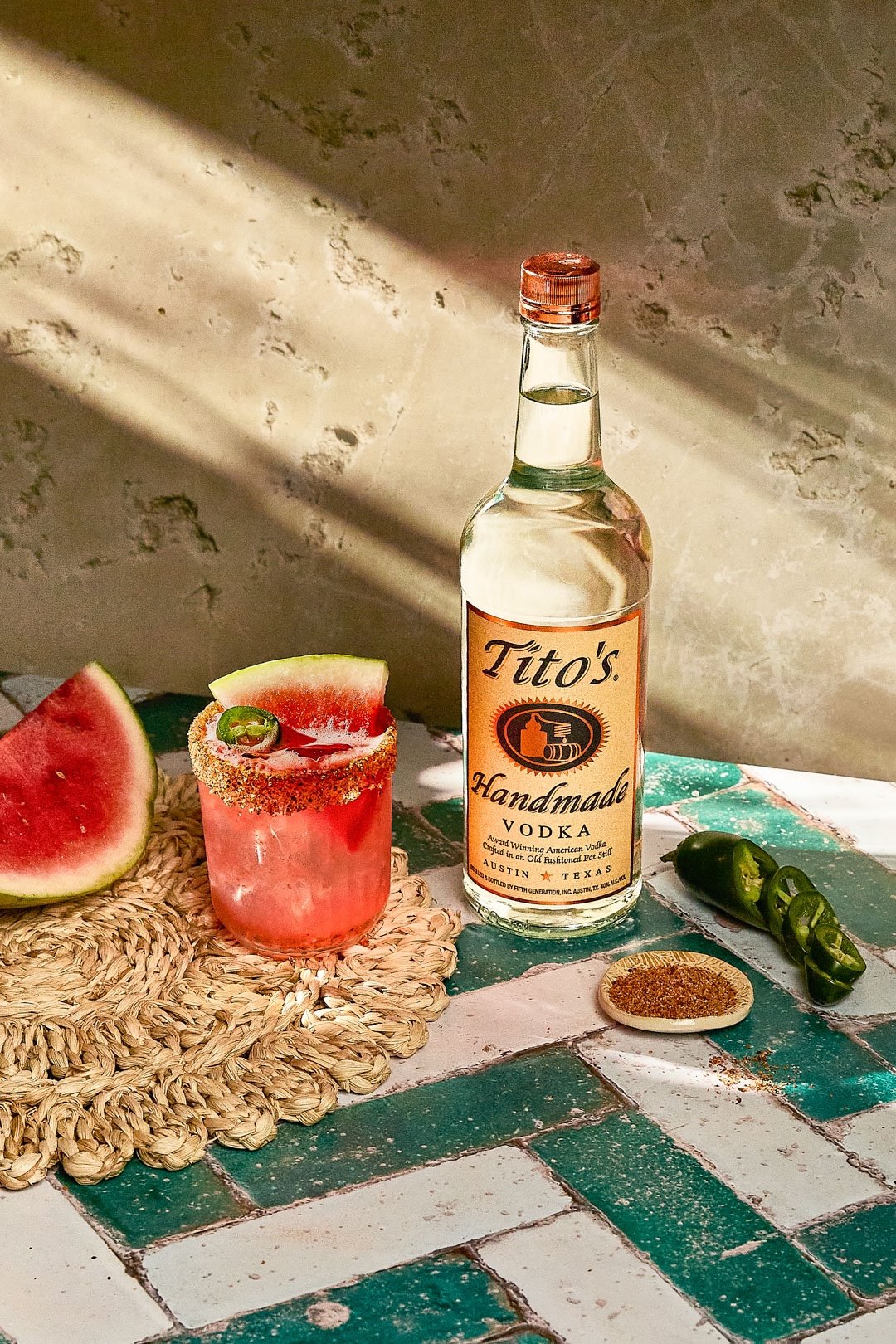 A bottle of Tito's Handmade Vodka, a watermelon cocktail garnished with watermelon and chili powder, a slice of watermelon, sliced jalapeño peppers, and a small dish of chili powder on a tiled surface with natural light.