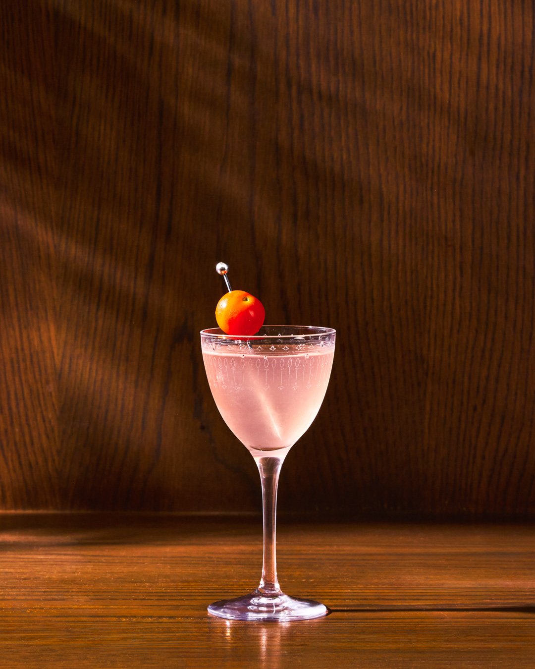 A pink cocktail in a stemmed glass garnished with a cherry tomato on a pick, placed against a dark wooden background.