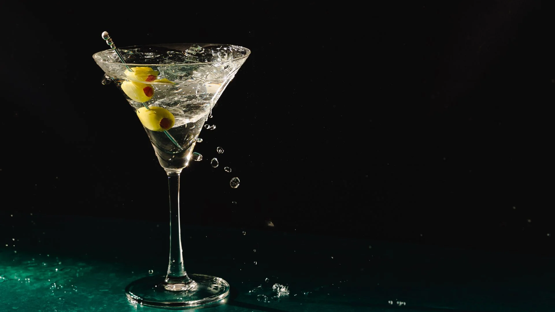 A martini glass with olives skewer and water splashing inside, against a black background.