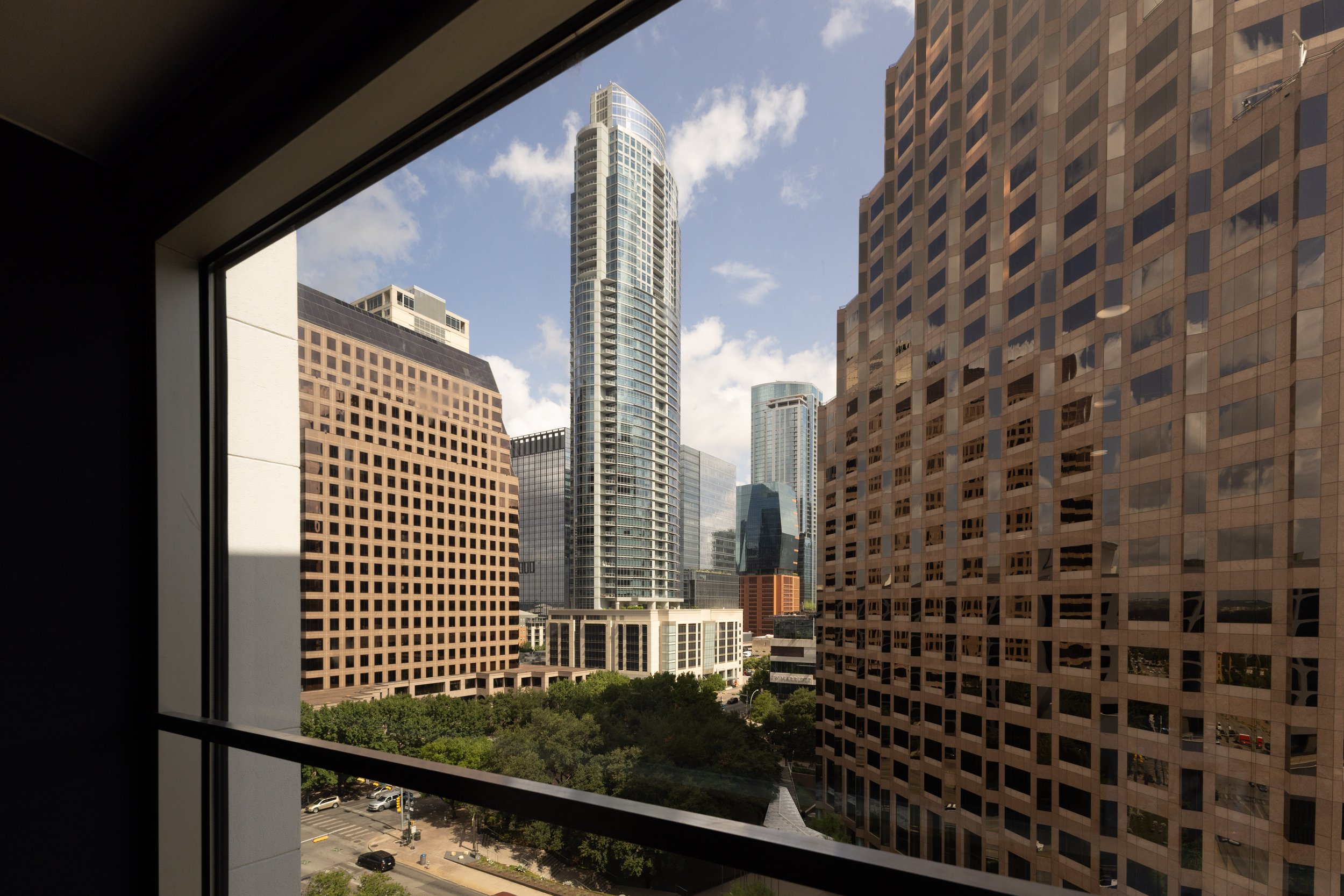 City skyline seen through a window, with tall modern buildings, trees, and a blue sky with some clouds.