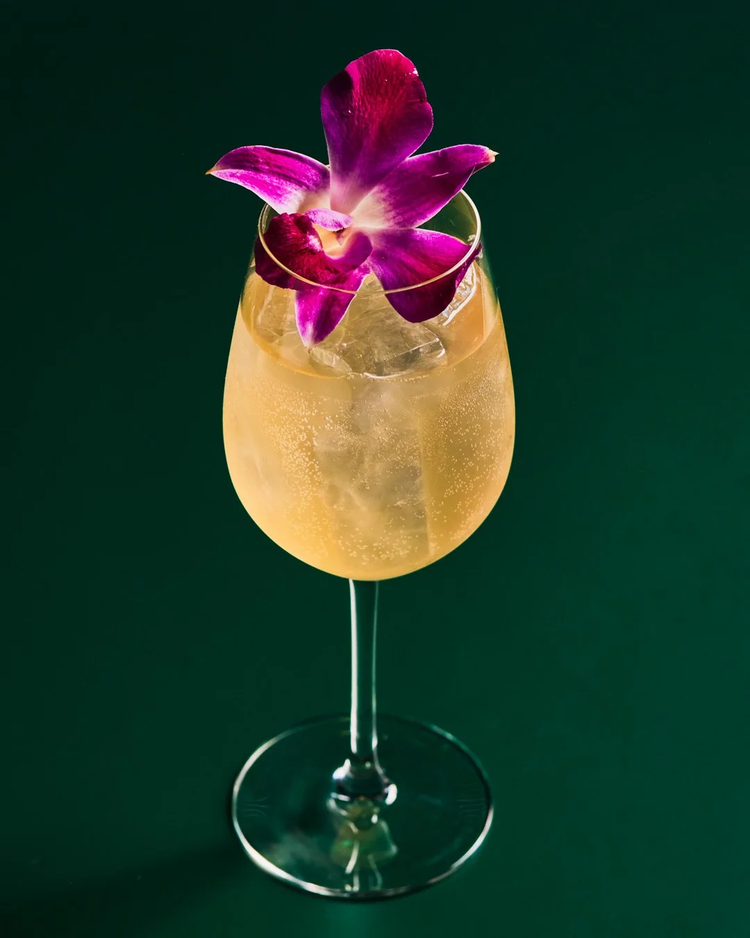 A cocktail in a stemmed glass with ice, garnished with a purple orchid flower, set against a dark green background.