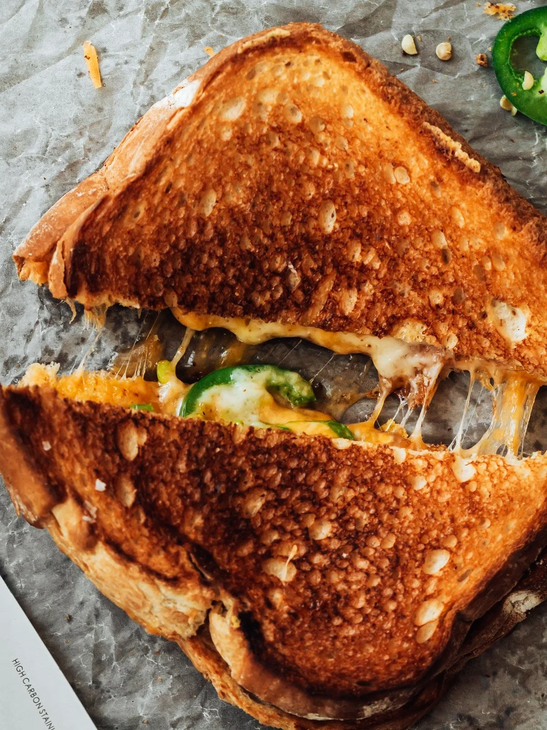 A close-up of a grilled cheese sandwich with melted cheese and jalapeno peppers on a piece of parchment paper.
