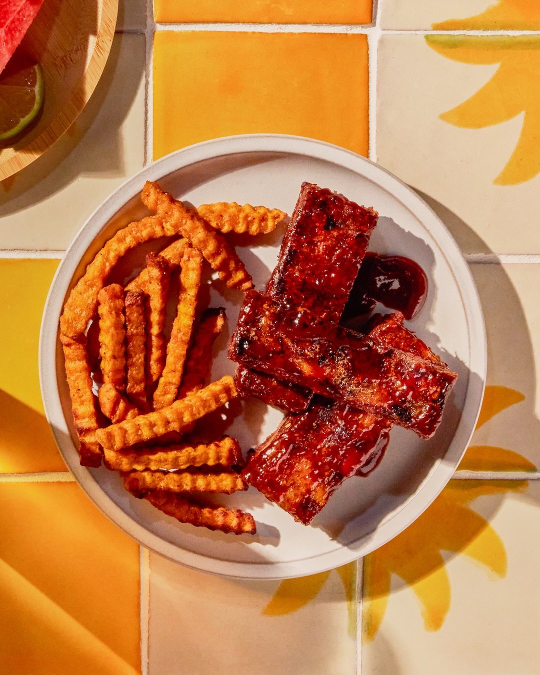 A white plate with crinkle-cut French fries and glazed pork ribs on a colorful table with yellow and orange tiles.