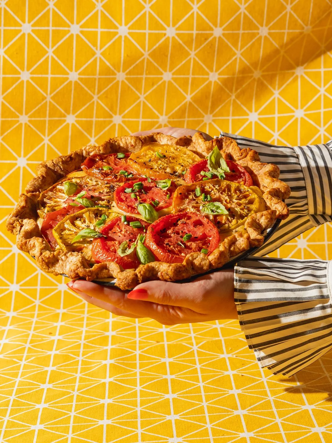 Someone holding a freshly baked vegetable quiche topped with sliced tomatoes and green herbs against a yellow patterned background.