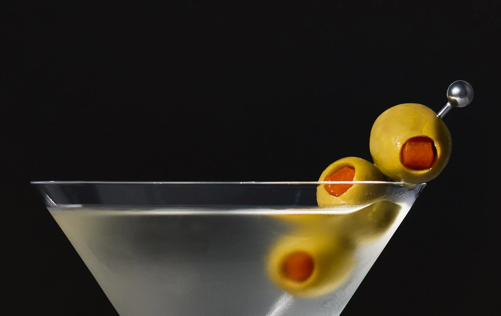 A martini glass filled with a clear liquid, garnished with three green olives, two with pimentos, skewered on a cocktail pick, set against a black background.