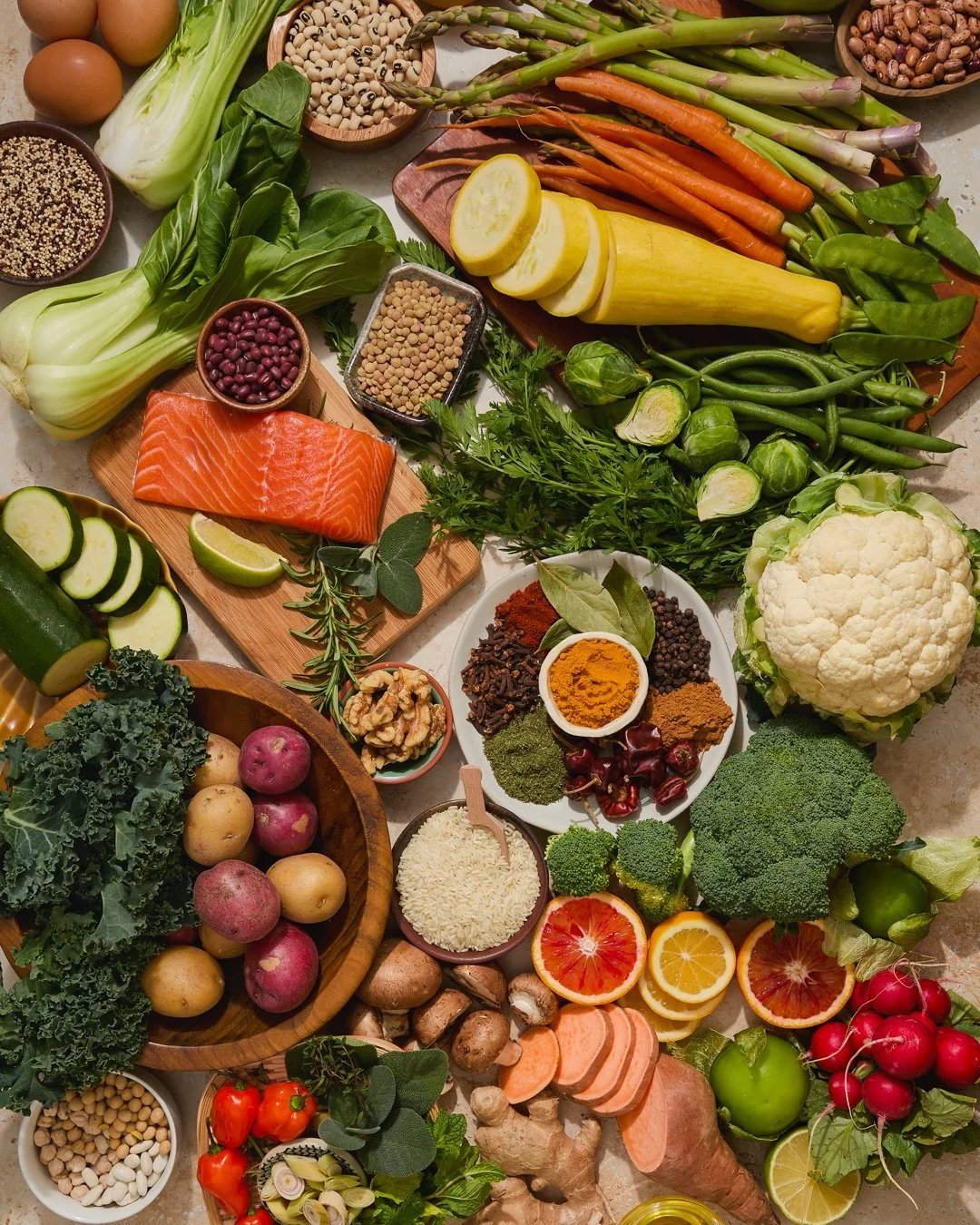 Assorted fresh vegetables, fruits, herbs, seeds, and a piece of raw salmon arranged on a table, including cauliflower, broccoli, carrots, radishes, lemon, blood and regular oranges, cucumber, zucchini, cherry tomatoes, cauliflower, red onion, herbs, 