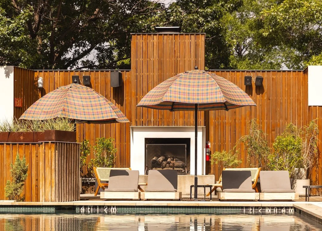 A poolside seating area with two large checkered umbrellas, chairs, and a white fireplace surrounded by a wooden wall and green plants.