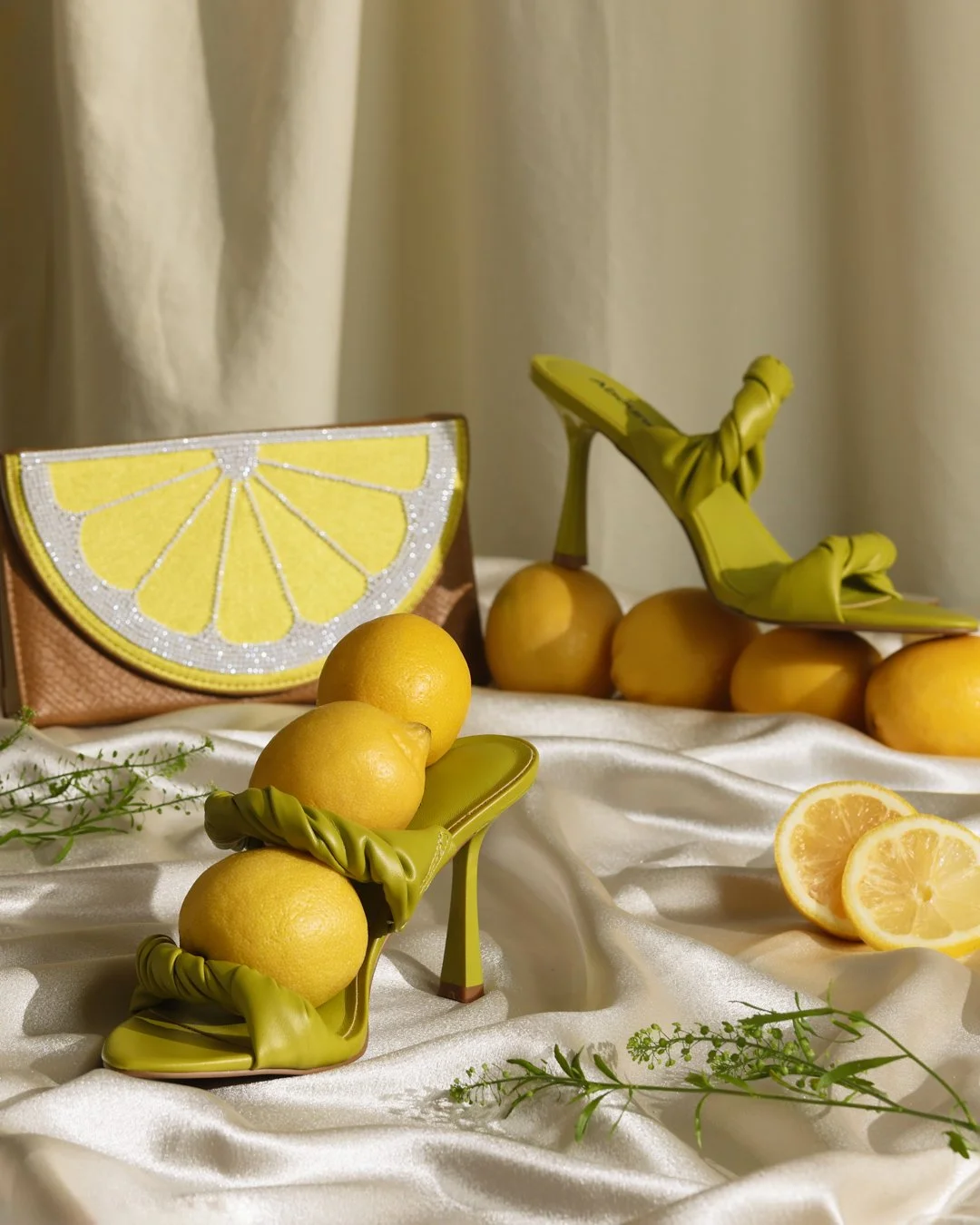 Table display with lemon-themed accessories, including yellow lemons, a lemon-shaped handbag, lemon-shaped high heels, and lemon slice decorations, set on a white satin cloth with green foliage.