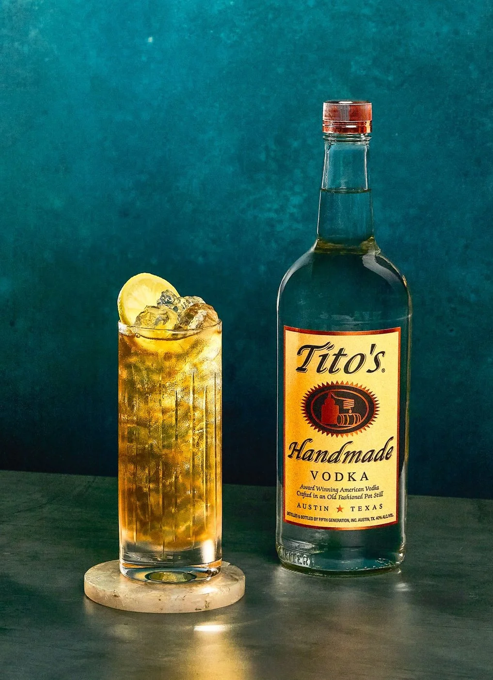 A tall glass of a golden-colored cocktail with ice and a lemon slice, next to a bottle of Tito's Handmade Vodka on a dark wooden surface.