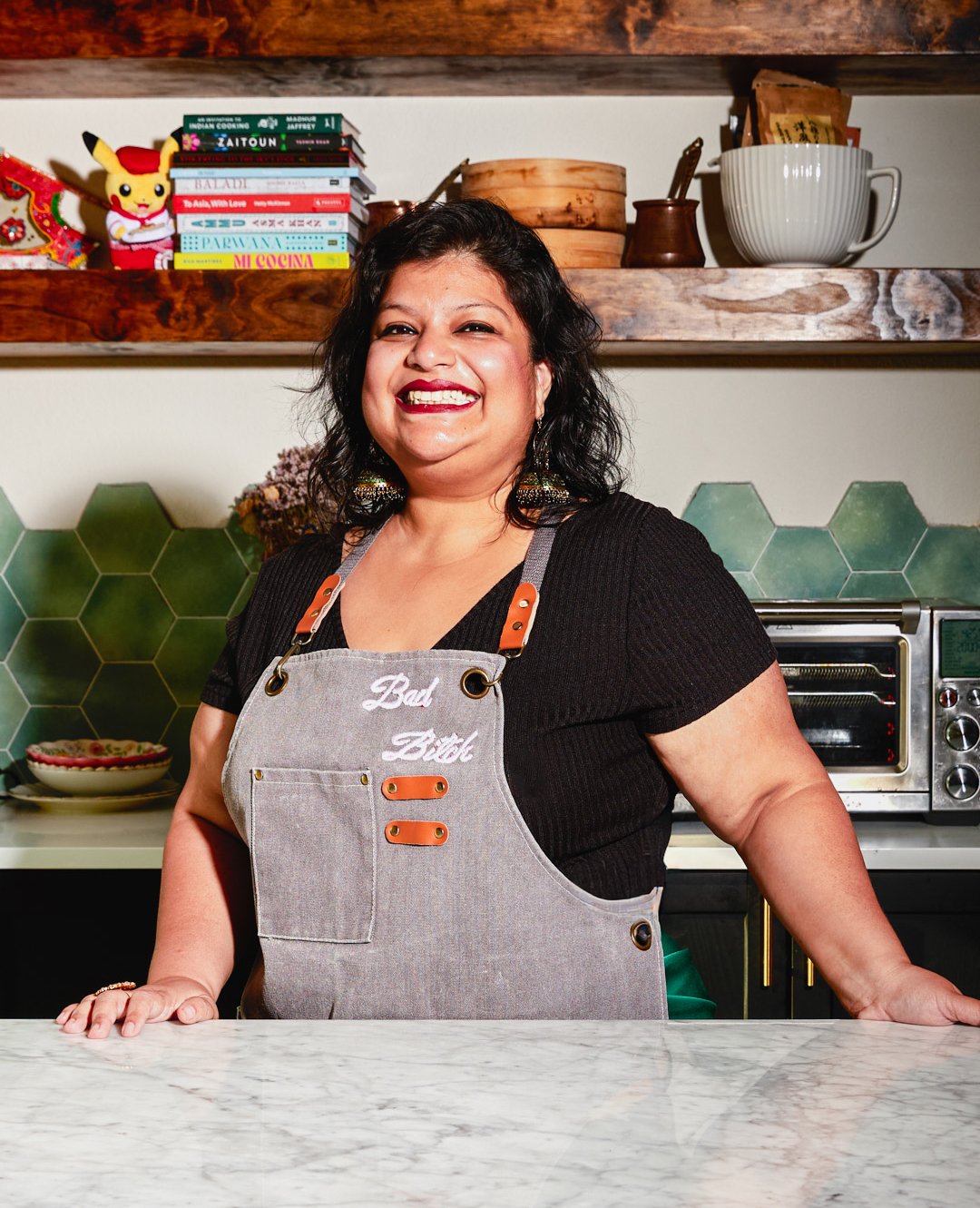 A woman smiling in a kitchen, wearing a black shirt and a gray apron with embroidery that says 'Bad Bitch'. She is standing behind a marble countertop, with green hexagonal tiles on the wall and shelves with cookbooks, decorative items, and kitchenwa