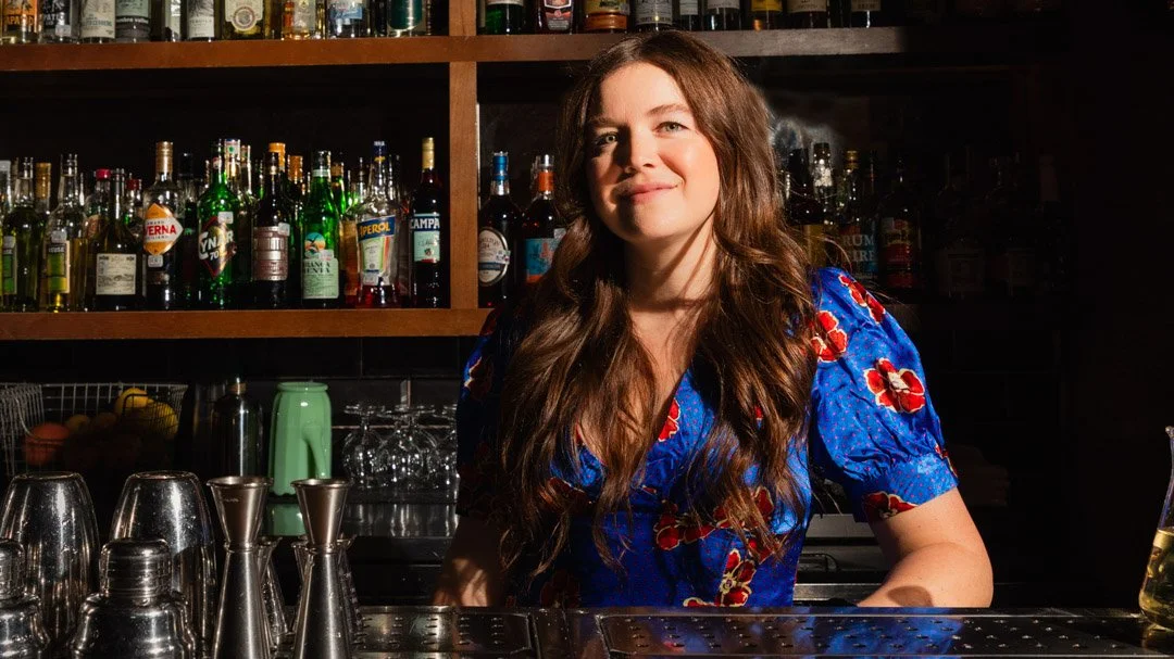 Woman behind a bar with liquor bottles on shelves, smiling and looking at the camera