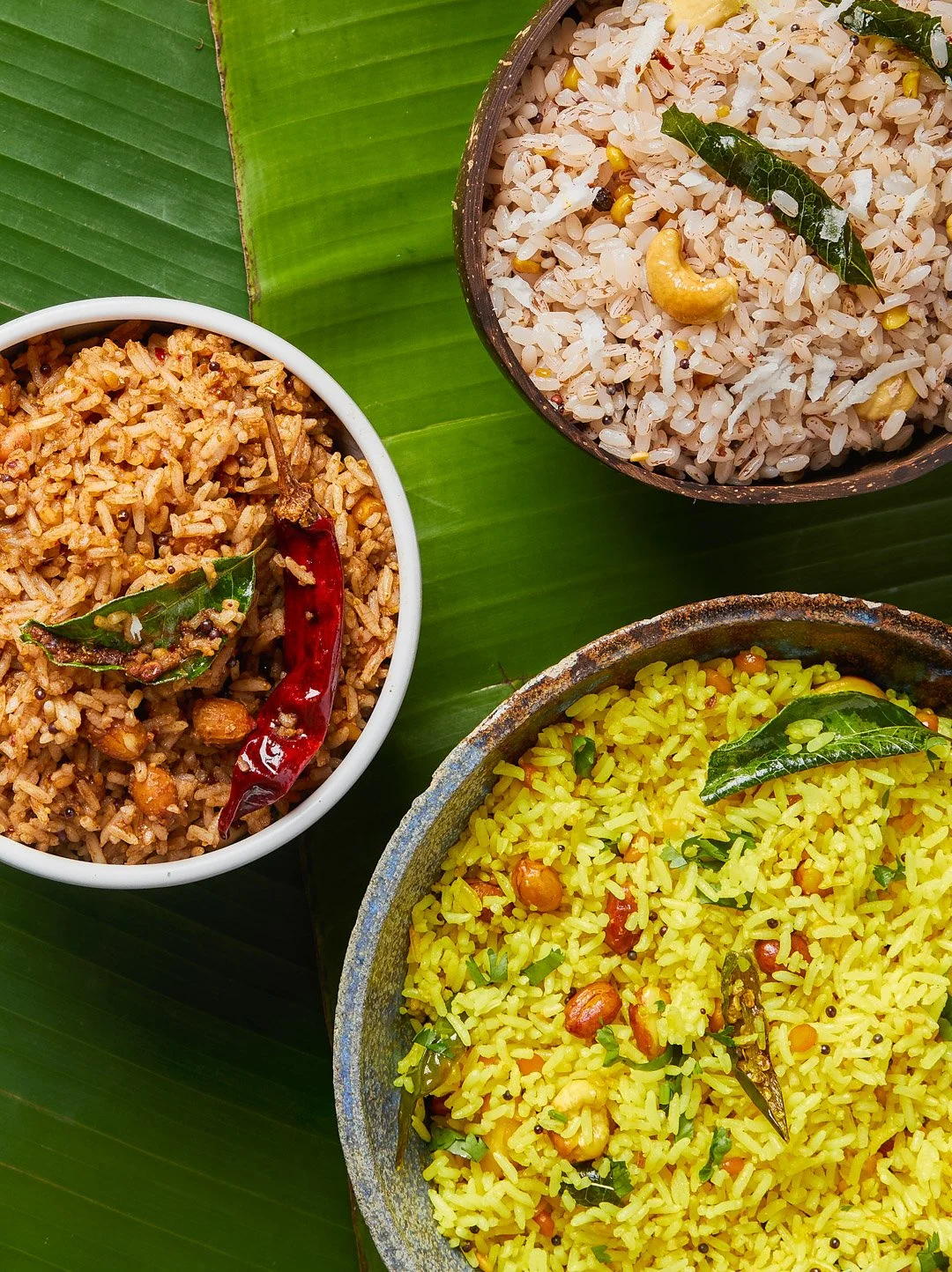 Three bowls of Indian rice dishes placed on a green banana leaf. The bowls contain different types of seasoned rice garnished with green curry leaves.