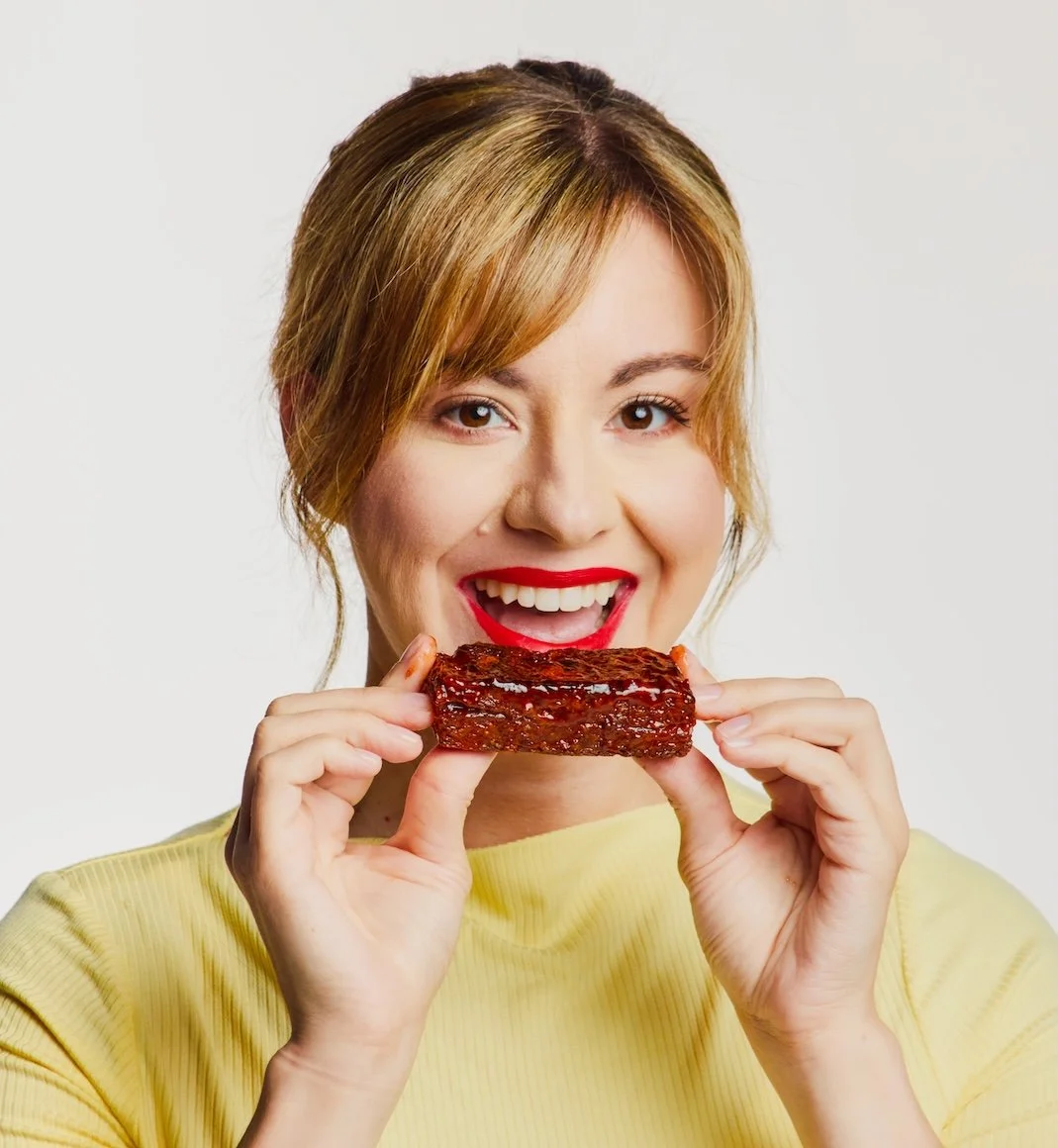 A woman with red hair and red lipstick holding a piece of glazed meat