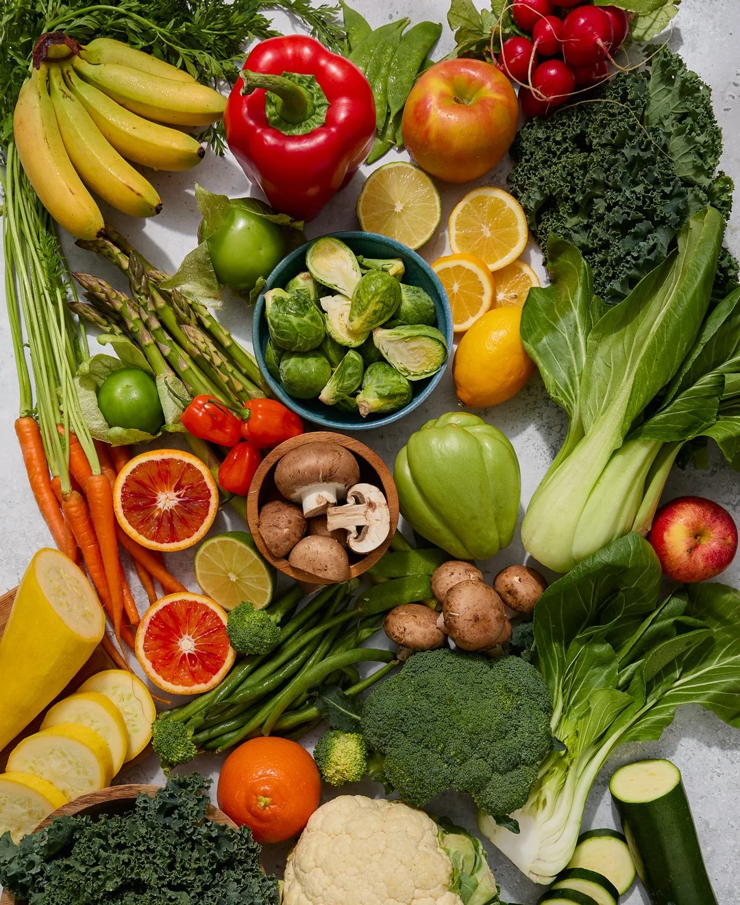 Variety of fresh fruits and vegetables including bananas, red bell pepper, apple, lemon, orange, radishes, kale, chard, carrots, blood orange, mushrooms, broccoli, cauliflower, zucchini, and other produce on a flat surface.