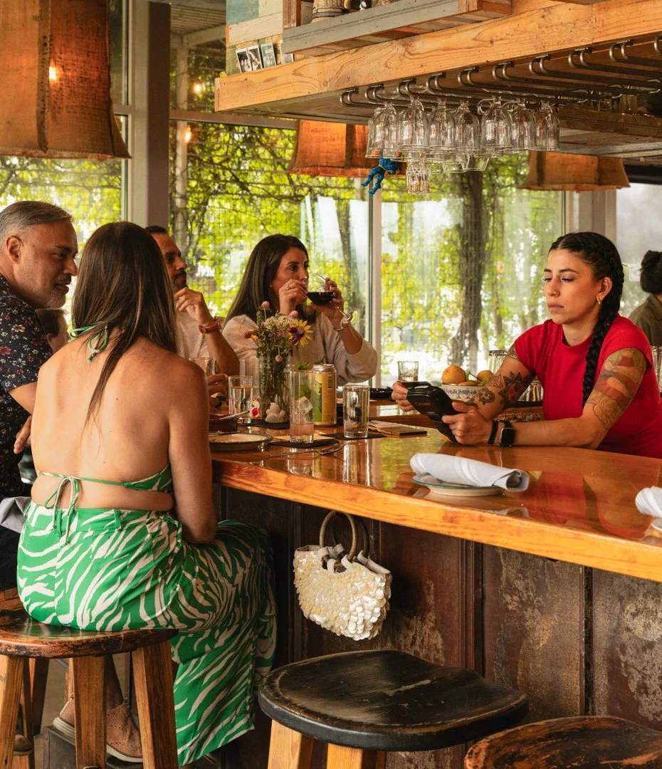 People sitting at a bar with one woman in a green dress and others drinking and talking while a bartender looks at her phone in a cozy, well-lit restaurant with large windows and greenery outside.