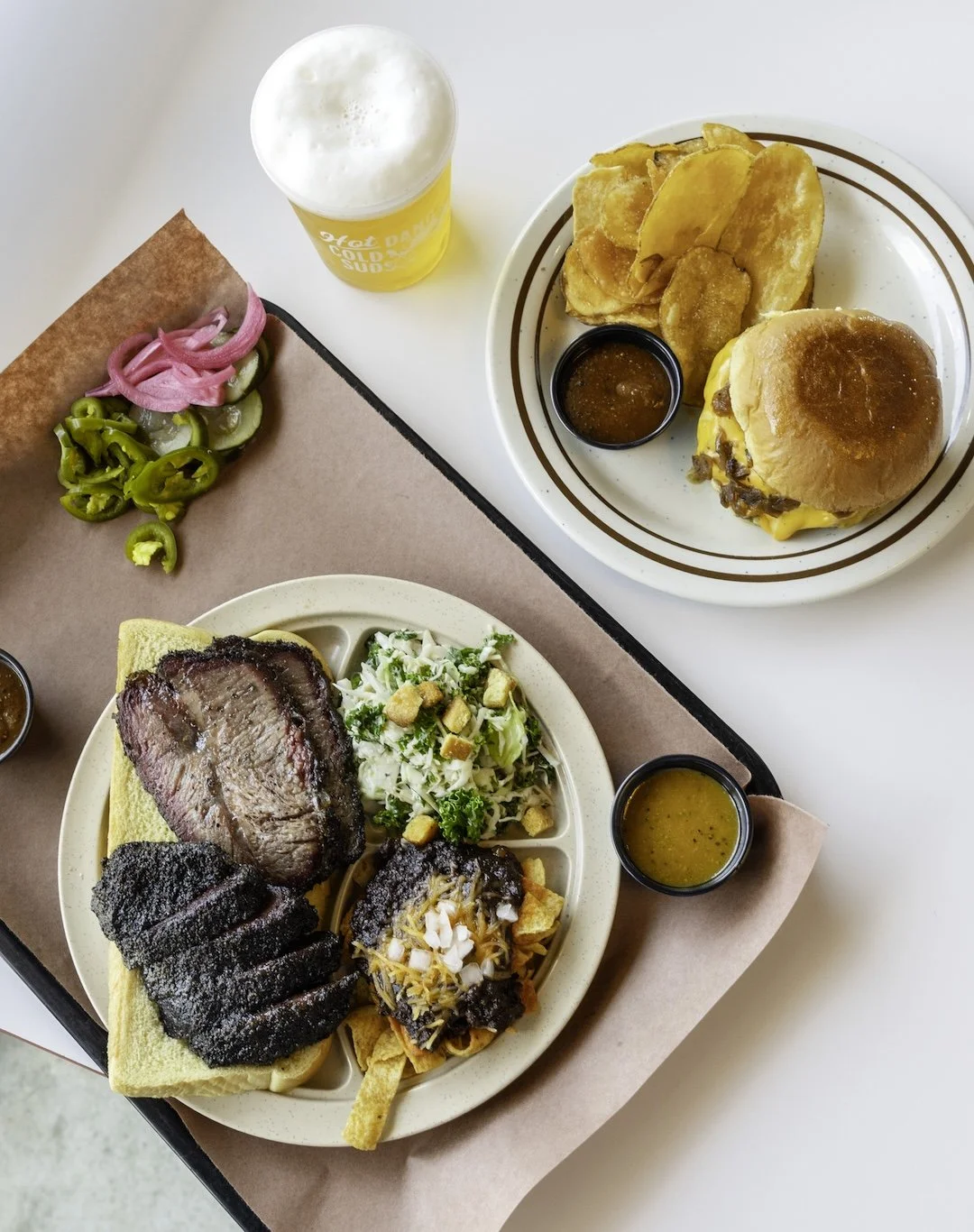 Meals including a cheeseburger with potato chips and dipping sauce, a plate with a sliced beef steak, BBQ pork, coleslaw, and pasta with sauce, a side of sliced pickles and onions, and a cup of beer.