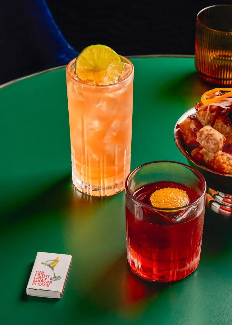 Two colorful cocktails on a green table with a bowl of food and a matchbox nearby.