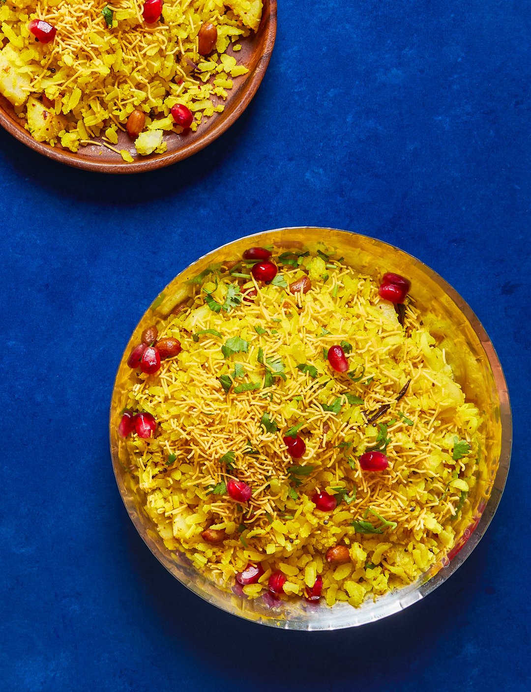 Close-up of a bowl of yellow Indian rice dish garnished with pomegranate seeds and chopped herbs, set on a blue surface, with a smaller bowl of the same dish nearby.
