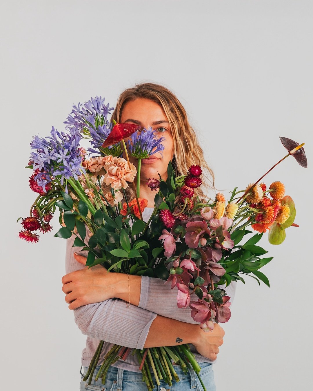 Woman holding large colorful bouquet of flowers, some covering part of her face, partially obscuring her expression, against a plain light gray background.