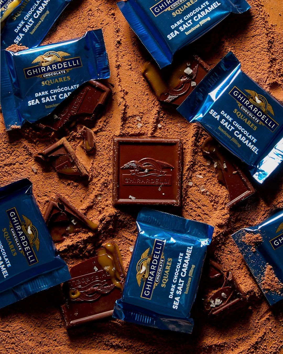 Assorted Ghirardelli dark chocolate squares with sea salt caramel flavor, placed on a surface dusted with cocoa powder, some partially unwrapped showing caramel and chocolate layers.