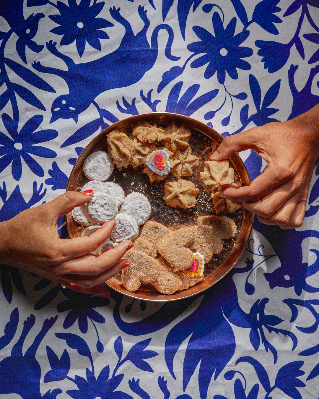 Hands holding a plate of traditional Mexican cookies on a blue and white floral tablecloth.