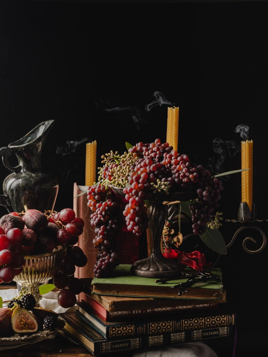 Still life of grapes, candles, books, and a silver pitcher against a dark background.
