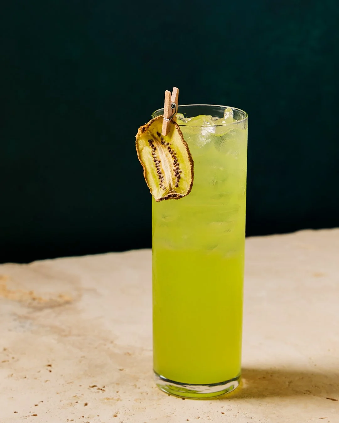 A tall highball glass filled with bright green kiwi-flavored cocktail and ice, garnished with a dried kiwi slice attached to a small clothespin.