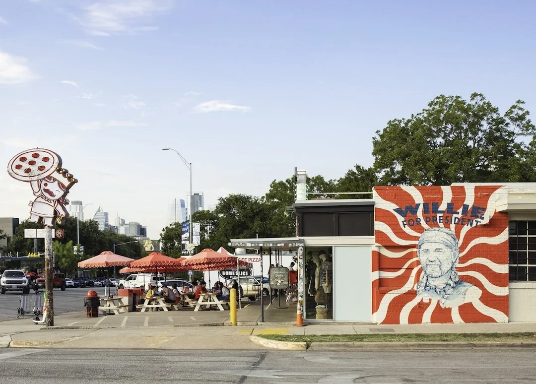 Sidewalk cafe with pink umbrellas, outdoor seating, and a mural of Willie Nelson on the wall.