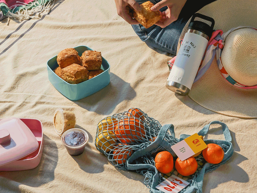 Picnic setup on a blanket with fried food, oranges in a mesh bag, a hat, water bottle, and condiments.