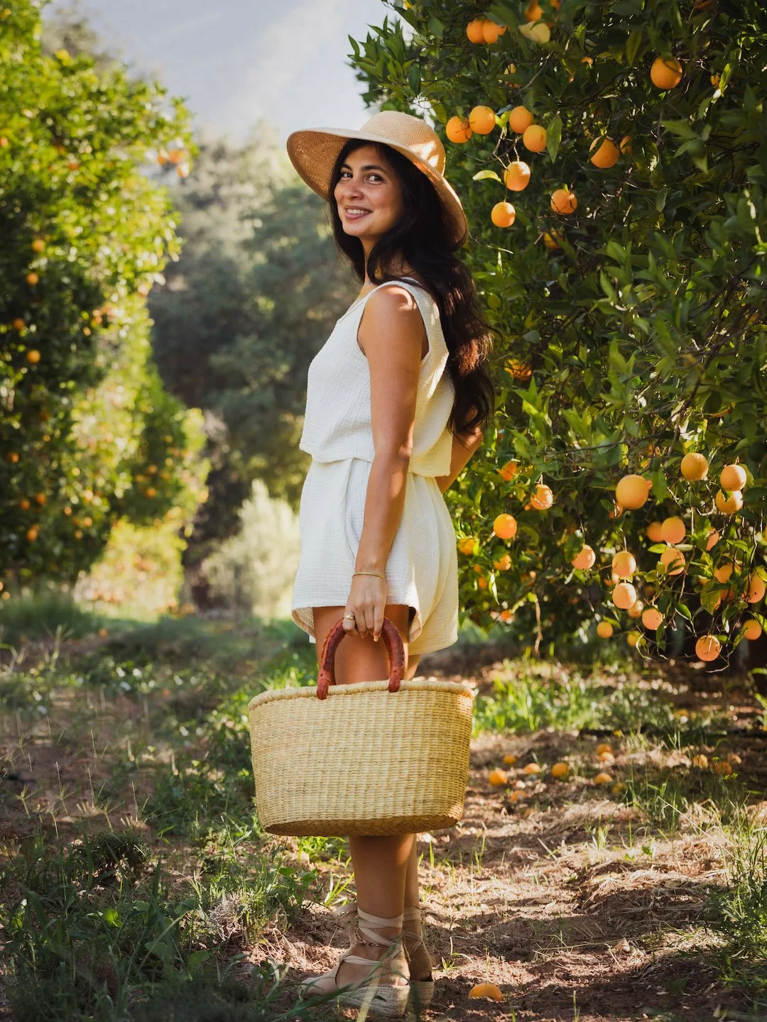 A woman in a white dress and sun hat is standing in an orange orchard, holding a woven basket, smiling, with orange trees in the background.