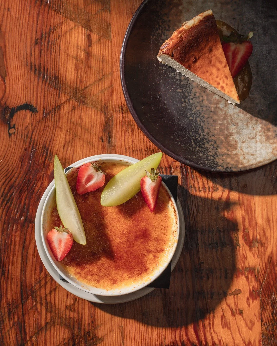 A dessert of crème brûlée topped with sliced strawberries and melon on a wooden table, with a slice of cake in a dark bowl.