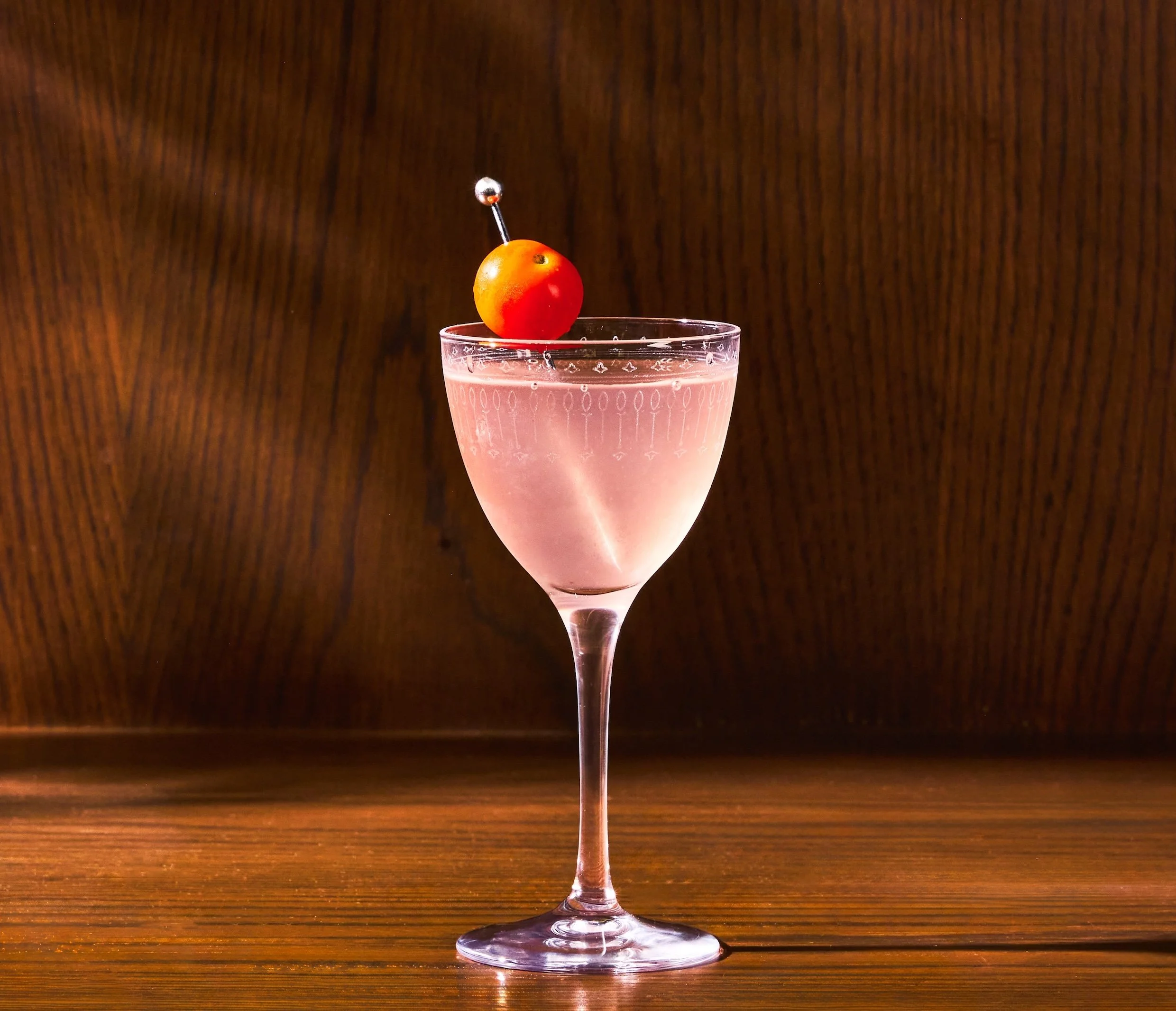 A pink cocktail in a stemmed glass garnished with a cherry tomato and a small metal pick, on a wooden table. Beverage photography by Nitya Jain for The Thompson Hotel Austin.