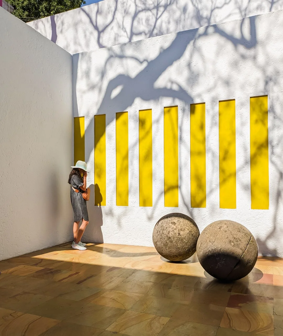A woman with a sunhat stands in front of a white wall with yellow vertical slats, shadows of tree branches are cast on the wall, and there are two large spherical rocks on the floor.